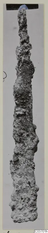 Long, narrow, heavily corroded iron tool photographed upright against a grey background, from Vouni