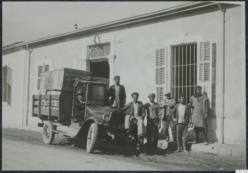 Lorry loaded with wooden crates parked outside a whitewashed building, with seven men posing beside it