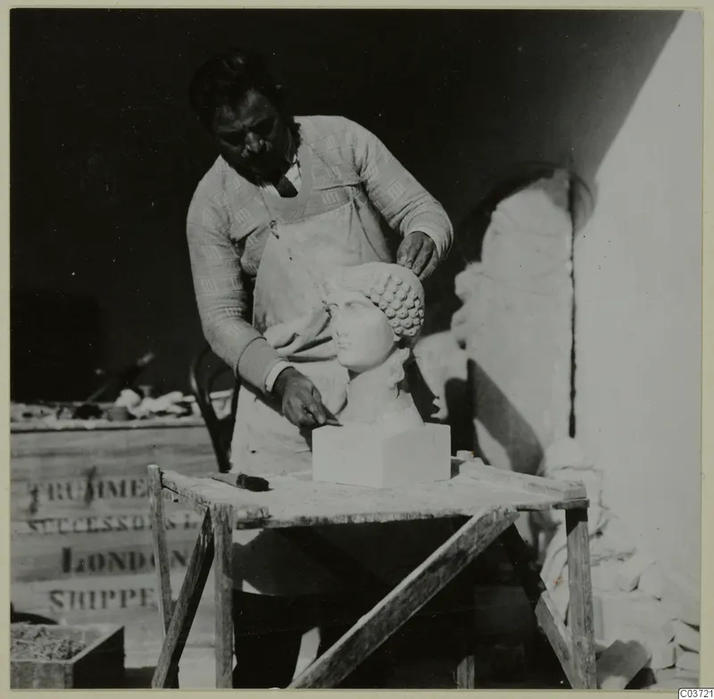 Person in an apron working on a pale sculpted female head on a wooden table, with a carved fragment behind