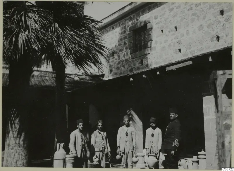 Five men stand with large clay vessels and baskets in a courtyard beside a stone building and palm trees