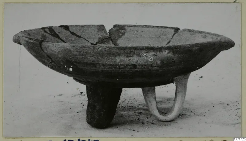 Pottery White Painted III-1 bowl on three legs, with a broken rim, standing on a dusty surface