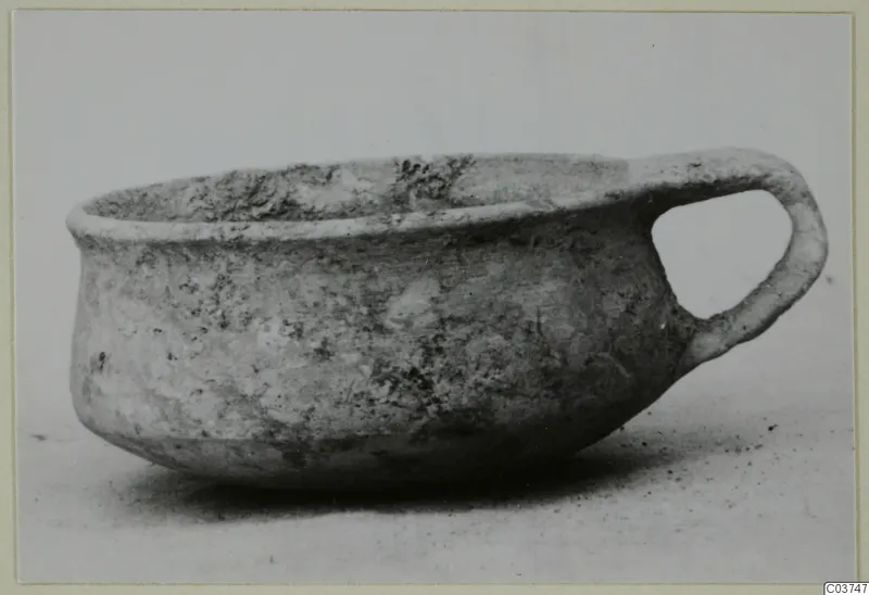 Plain white wheel-made bowl with a single loop handle, seen from the side.