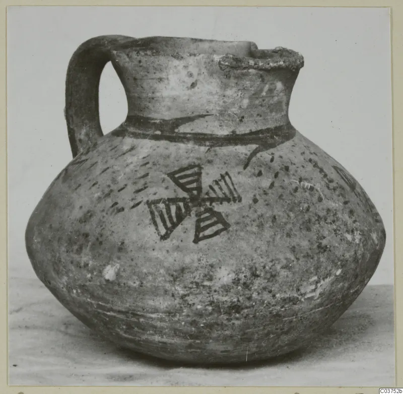Round-bodied jug with a strap handle and dark painted bands and a cross-like motif, photographed on a plain surface.