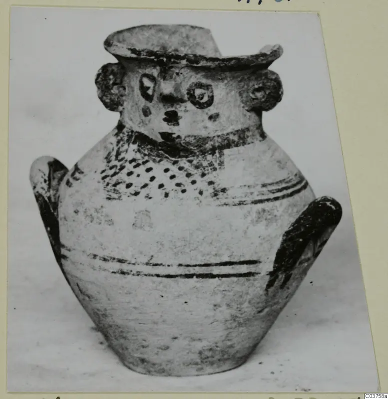 Pottery jug with a modelled face on the neck and painted dark bands, shown on a card mount