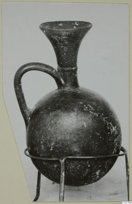 Dark-surfaced jug with a flaring neck and single strap handle, shown on a three-legged wire stand.