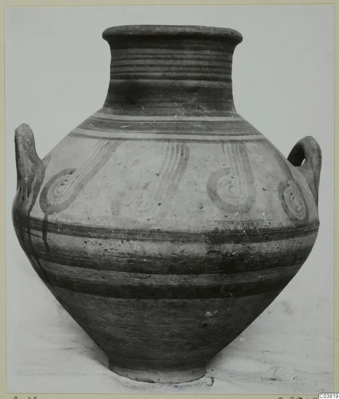 Two-handled painted jar with a flared rim, dark bands and dark spiral motifs on a pale ground, against a plain backdrop.