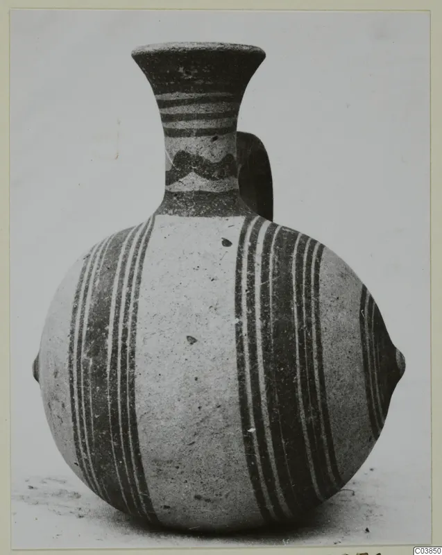 Painted pottery vessel with a flared neck, a loop handle and dark vertical bands on its rounded body, photographed against a grey backdrop.