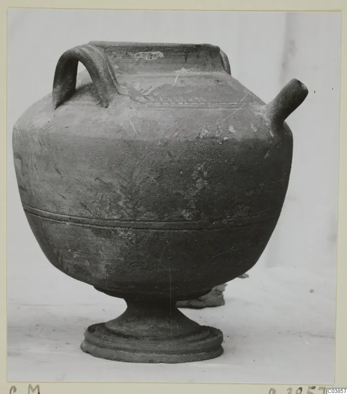 Pottery vessel with a loop handle, a tubular spout and a pedestal foot, photographed from the side.