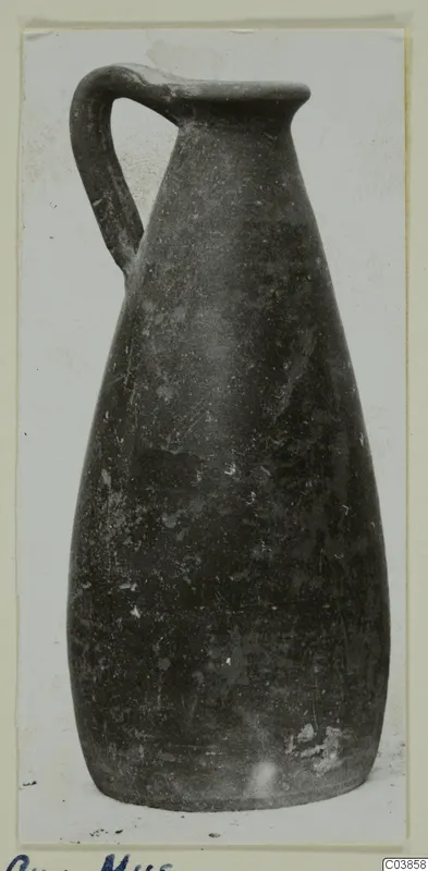 Tall dark jug with a single loop handle and a flared rim, photographed on a card mount
