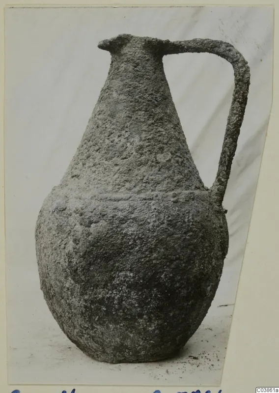 Corroded bronze vessel with a long neck and a single strap handle, shown against a light cloth backdrop
