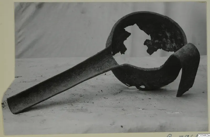 Corroded metal ladle with a long flat handle and a broken bowl, shown on a light surface.