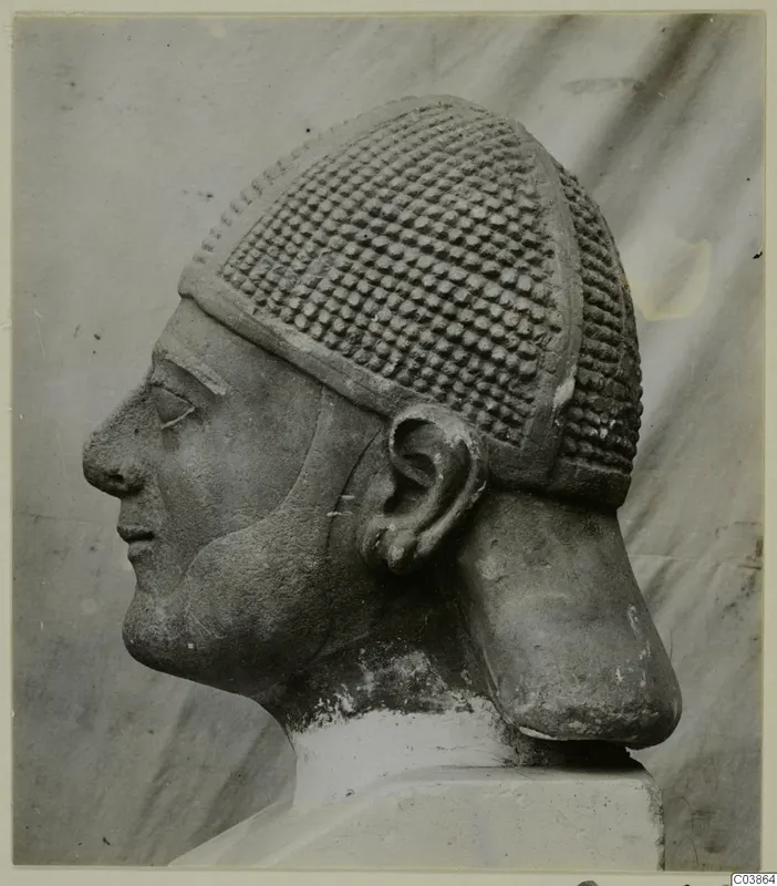 Limestone head in profile, wearing a cap covered in rows of raised knobs, on a plain base.