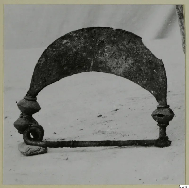 Corroded metal object with an arched plate, two knobbed uprights and a horizontal bar ending in a loop