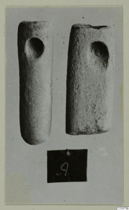Two cylindrical stone objects, each with a round hole near one end, photographed on grey card with a number label below.