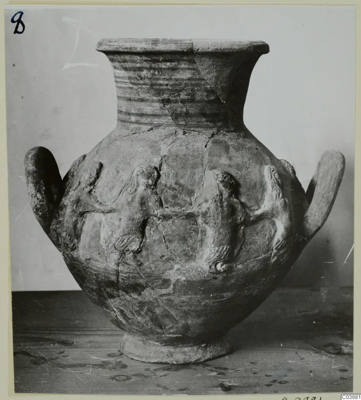 Pottery jar with a flared neck, two handles and raised relief figures on the body, standing on a wooden table