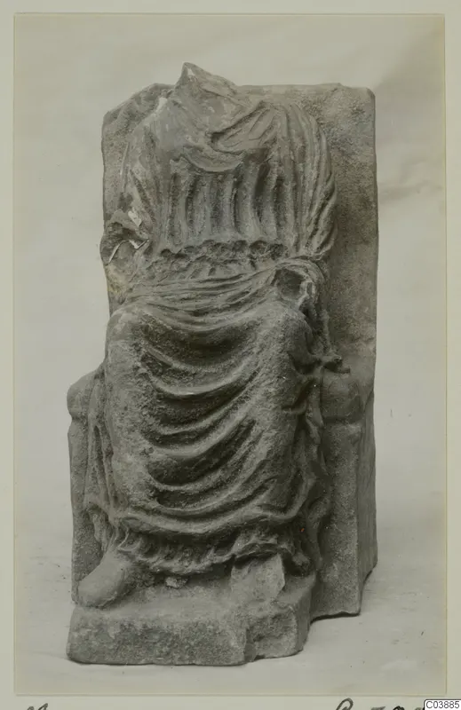 Carved stone fragment of a draped figure with a damaged head, on a rectangular block.