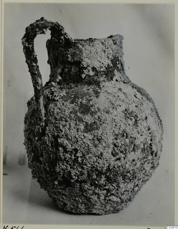 Corroded bronze jug with a looped handle, photographed against a plain grey background.