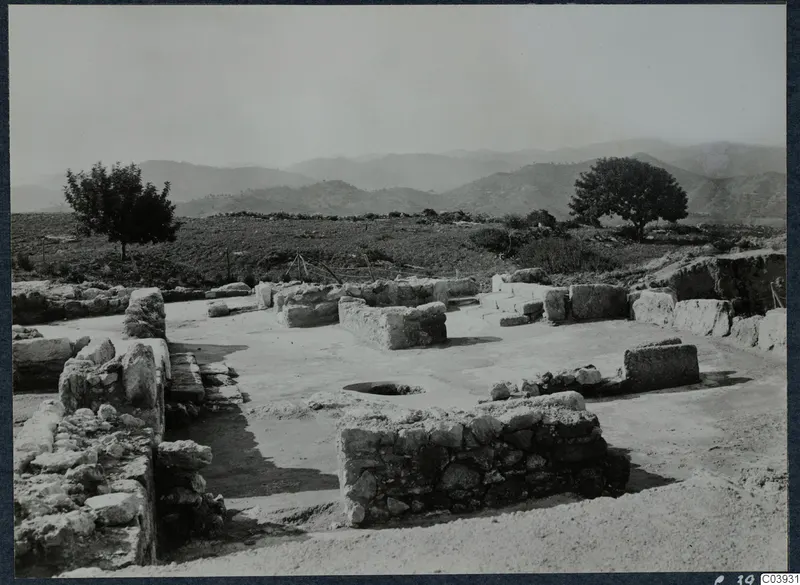 Ruined stone walls and a round hole in a flat paved area at Vouni palace, with trees and hills behind.