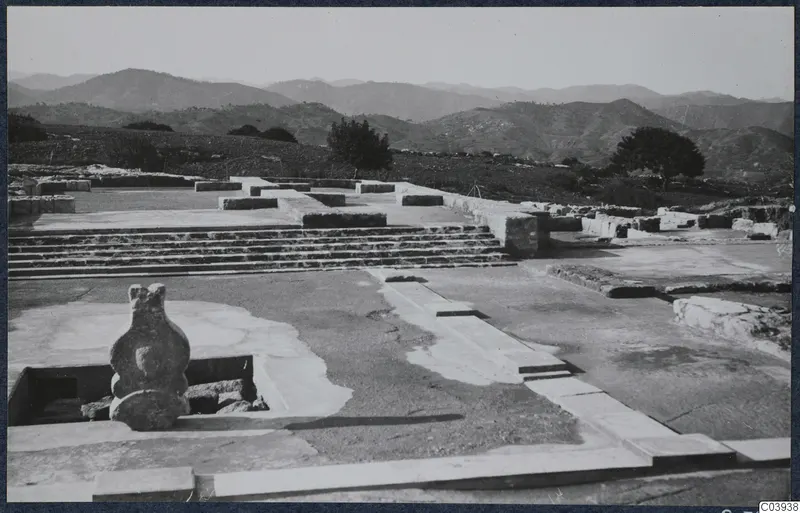 Stone steps and low wall foundations of the palace ruins at Vouni, with a stone figure in the foreground and hills behind.