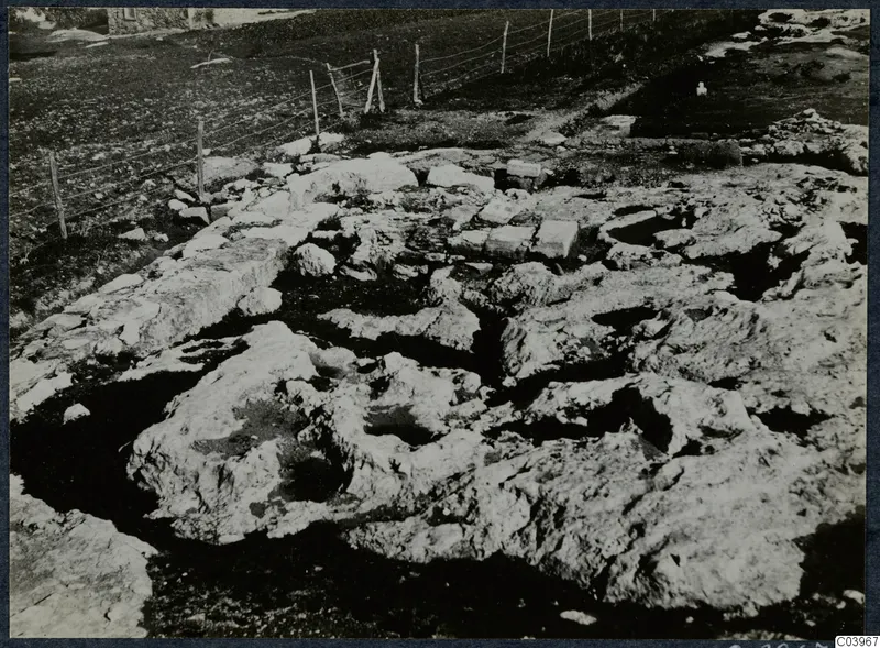 Stone foundations and cut bedrock at Vouni, with a wire fence and a stone building beyond.