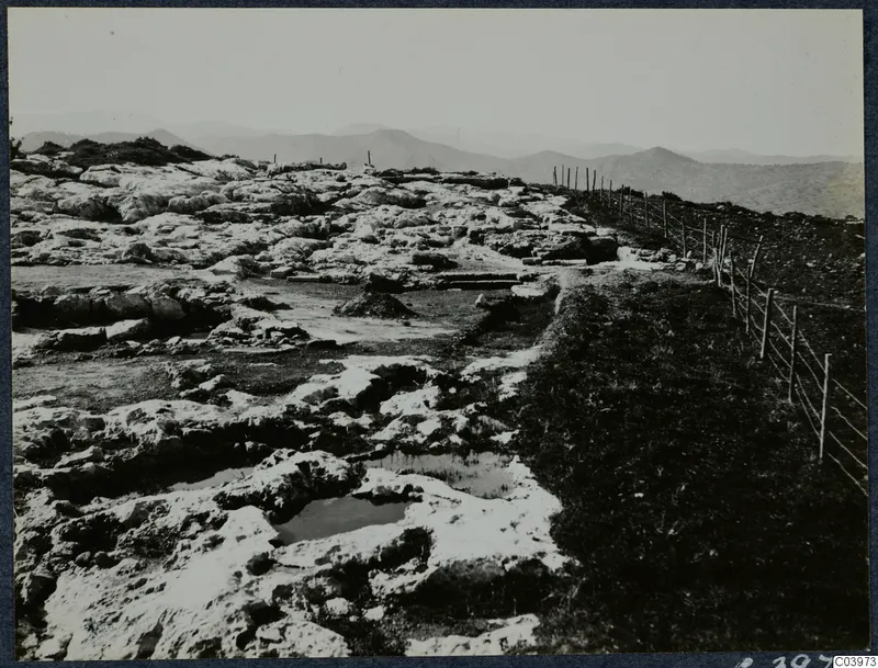 Pale rock ground at Vouni with cut channels and shallow pools, a wire fence on the right and hills behind