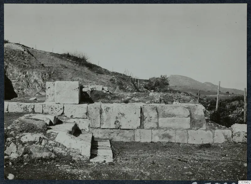 Large dressed limestone blocks of a wall and a stepped structure at Soli, with a hillside and wire fence behind.