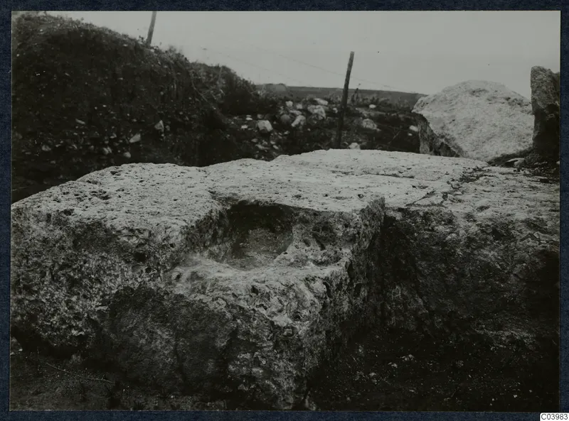 Large pitted stone block with a rectangular hollow in its top, at the theatre at Soli, with a fence post and stones behind.