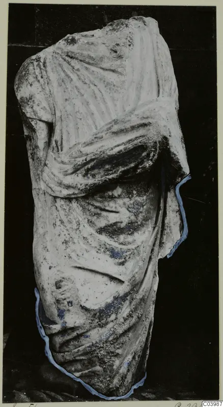 Marble statue fragment of a draped figure, headless, with one arm bent across the body.
