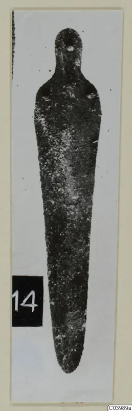 Bronze tool with a loop at one end and a long tapering blade, from Lapithos, shown upright on a white card