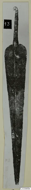 Corroded metal blade with a curled loop at the top, from Lapithos, photographed on a pale card strip.