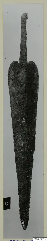 Corroded metal blade with a narrow tang, photographed upright against a grey background, from Lapithos