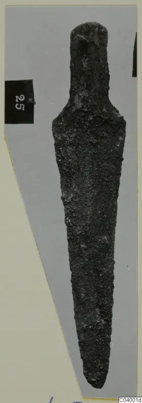 Corroded bronze blade-shaped tool from Lapithos, with a narrow tang, photographed on a grey card.