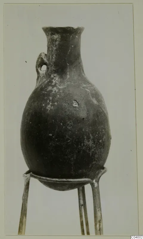 Red Polished III jug from Lapithos, standing on a three-legged wire stand against a plain background