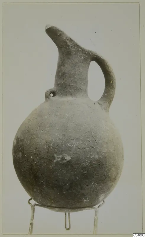 Pottery Red Polished III jug with a round body, tall neck, spout and loop handle, shown on a wire stand