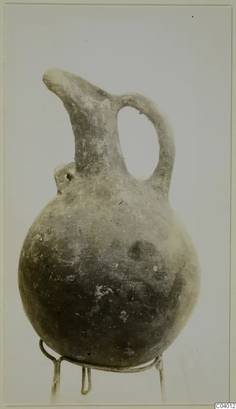 Red polished jug with a single loop handle and a pinched spout, shown on a wire stand against a plain background