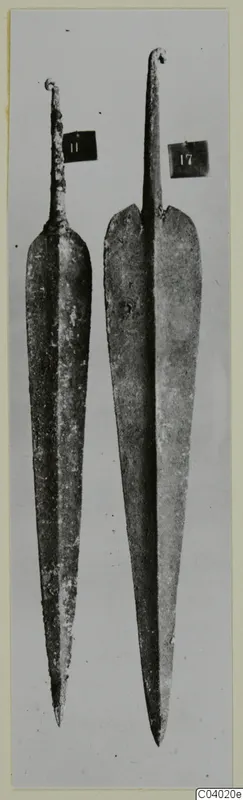 Two long metal blades with narrow tangs, photographed upright side by side against a grey background