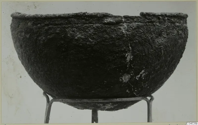 Deep rounded bowl with a dark, encrusted surface and broken rim, standing on a metal wire stand