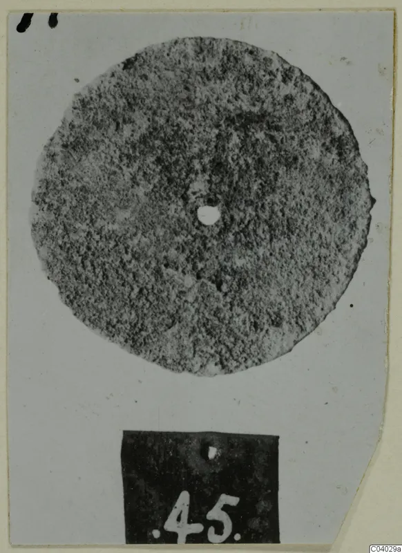 Corroded bronze disc with a central hole, photographed on a card with a number label below it.