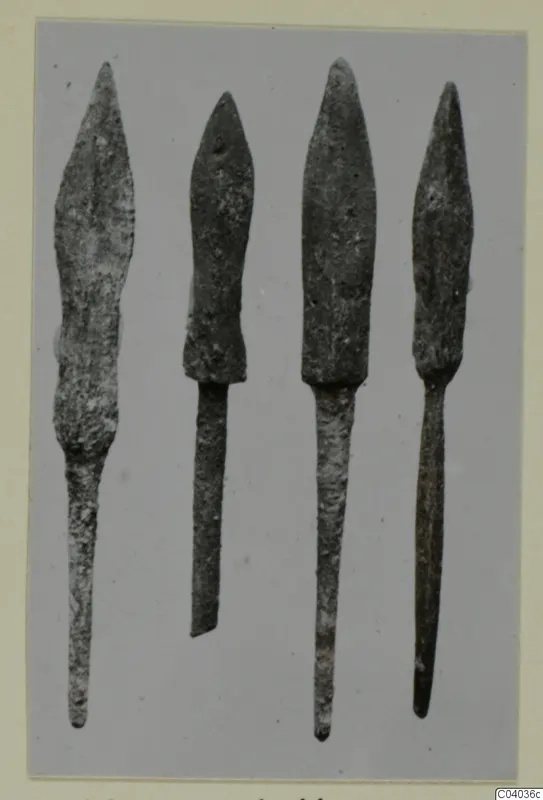 Four pointed metal blades with long tapering tangs, standing side by side against a grey background.