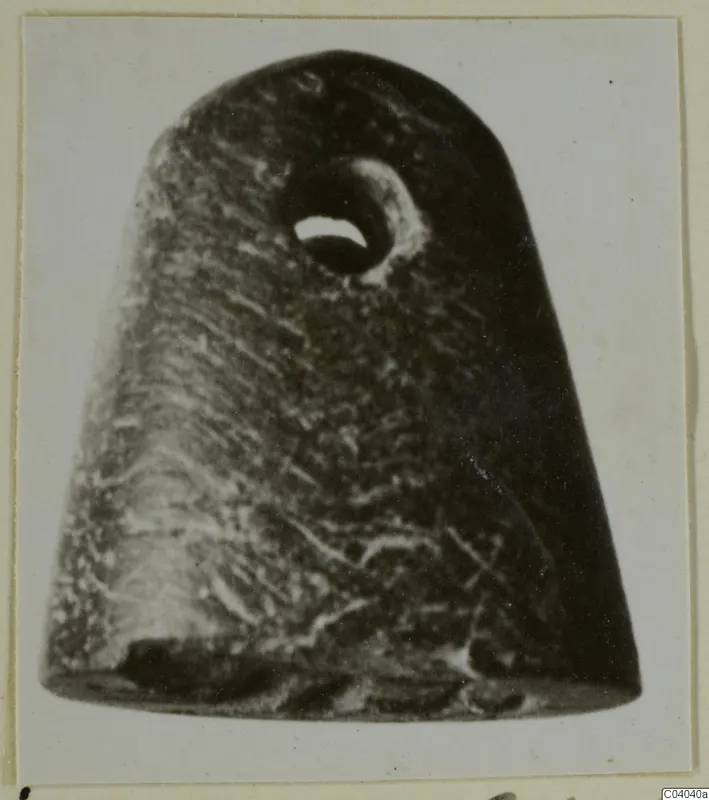 Dark stone object with a round perforation near the top, photographed on a pale card mount