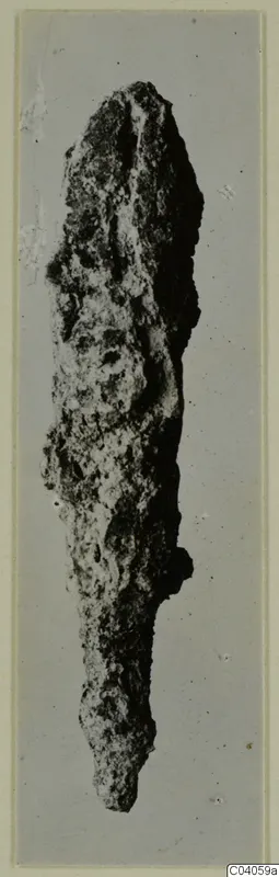 Heavily corroded iron object, long and narrow, photographed upright against a plain grey backdrop