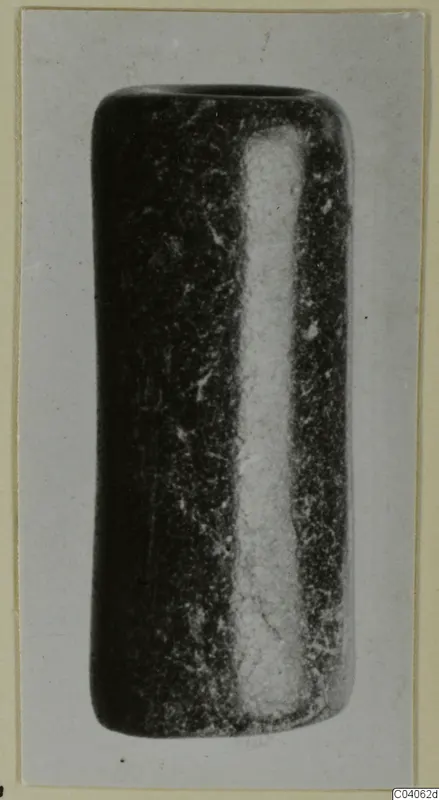 Dark stone object shaped like a short cylinder with a pale vertical band, shown on a grey card mount