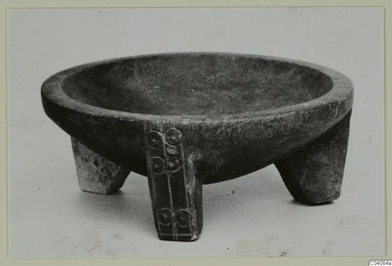 Stone bowl standing on three short legs, one leg with a band of circular motifs carved down it.