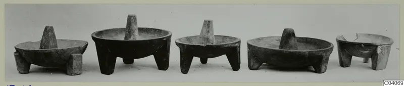 Five dark stone bowls on short legs, each with a central upright pestle, set in a row against a plain background
