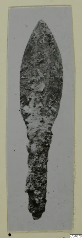 Corroded metal blade with a leaf-shaped tip and a long narrow tang, shown upright on a card mount