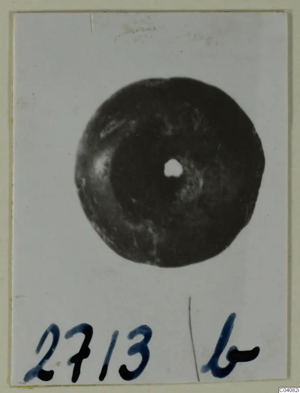 Round dark object with a central hole, photographed face-on on a white card with a handwritten number label.
