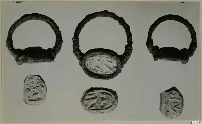 Three silver rings with oval bezels above three engraved glyptic objects, photographed on a grey background