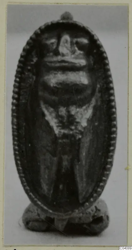 Small oval pendant with a beaded rim and a face-like relief, shown against a grey backdrop.