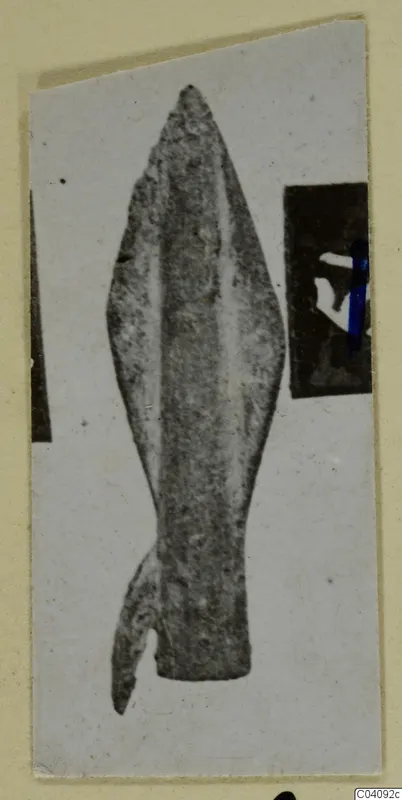 Bronze weapon head, leaf-shaped with a central ridge and a broken lower end, photographed on a mounted card
