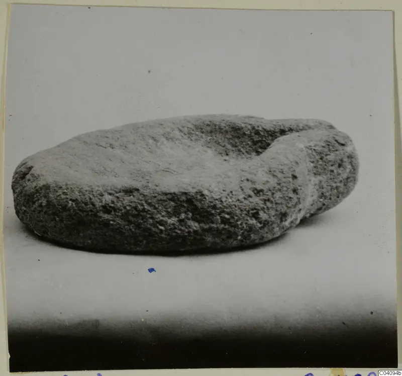 Rough stone object with a shallow hollow on top, photographed on a pale surface.